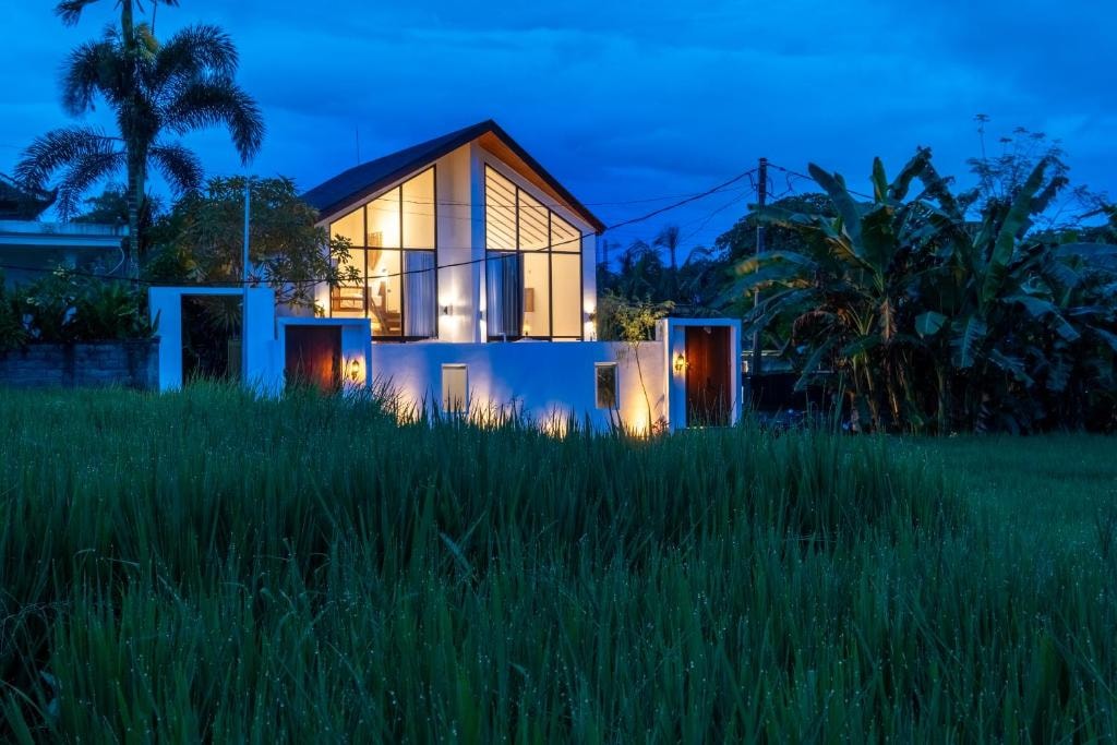 The Santa Ubud Resort at dusk, its gabled two-storey villa lit from within and seen across the rice field