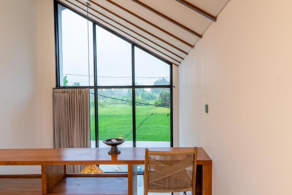 Timber desk and woven chair beneath a tall gable window looking out over a bright green rice field